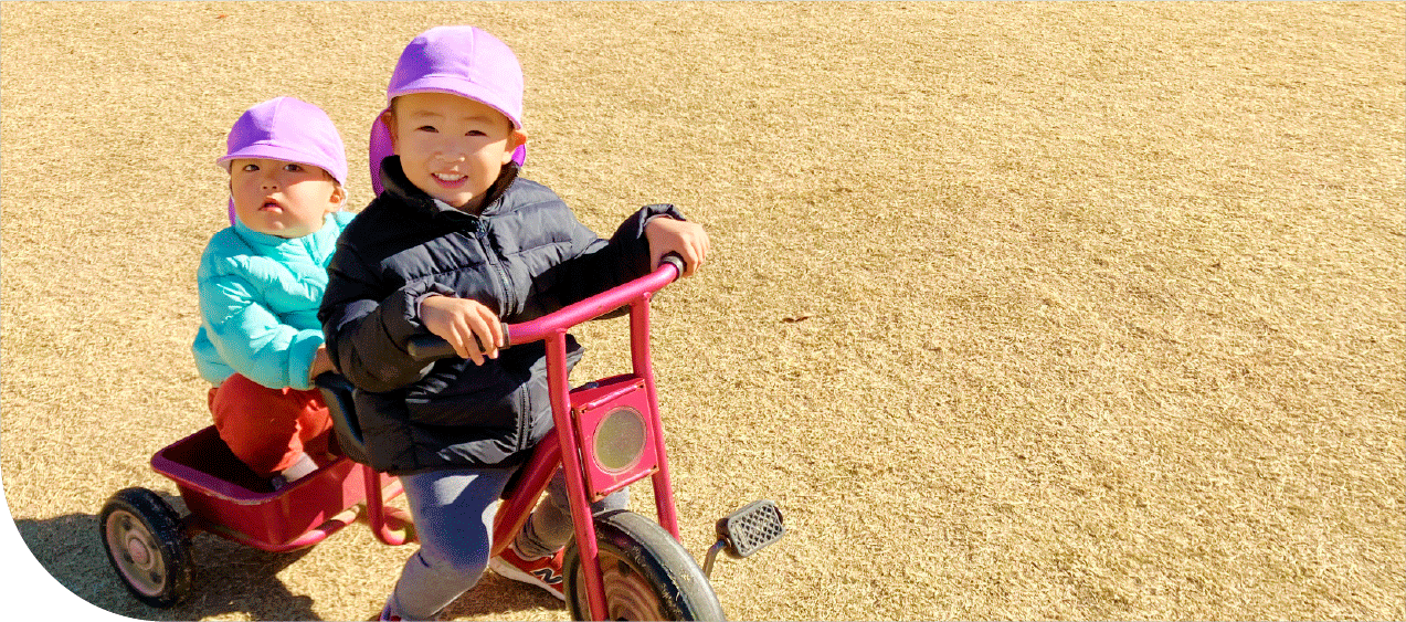 公園で三輪車の遊具に乗っている園児2名。1名は荷台のようなスペースに乗っている。